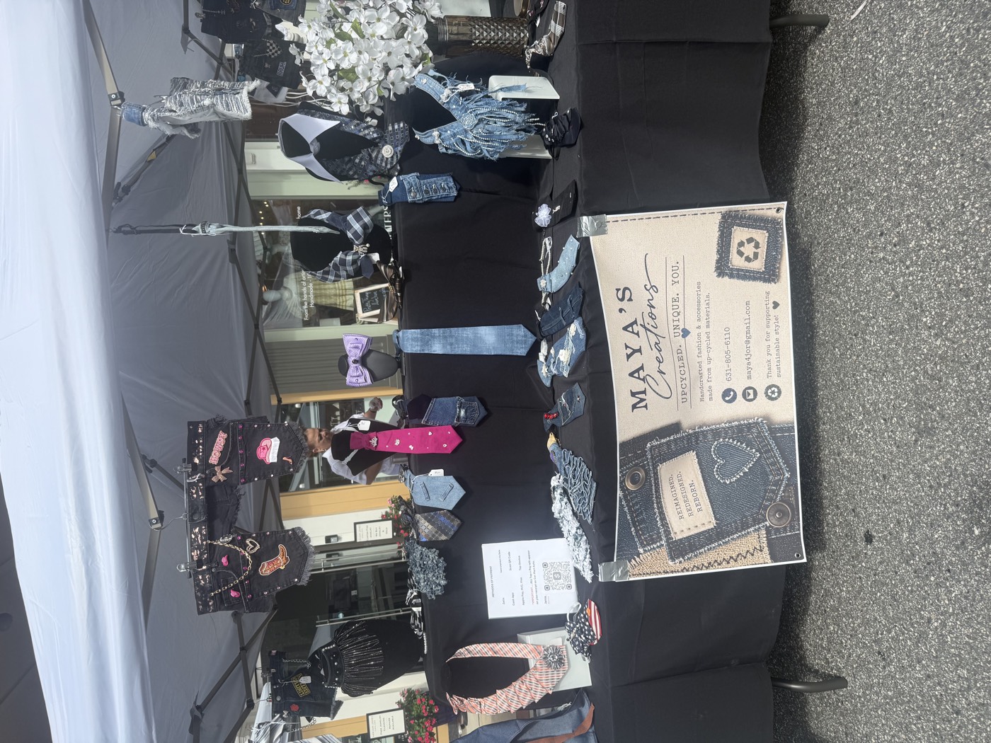 The Maya's Creations market booth: necktie necklaces, denim jewelry and bags on a black tablecloth, with the banner hanging in front.