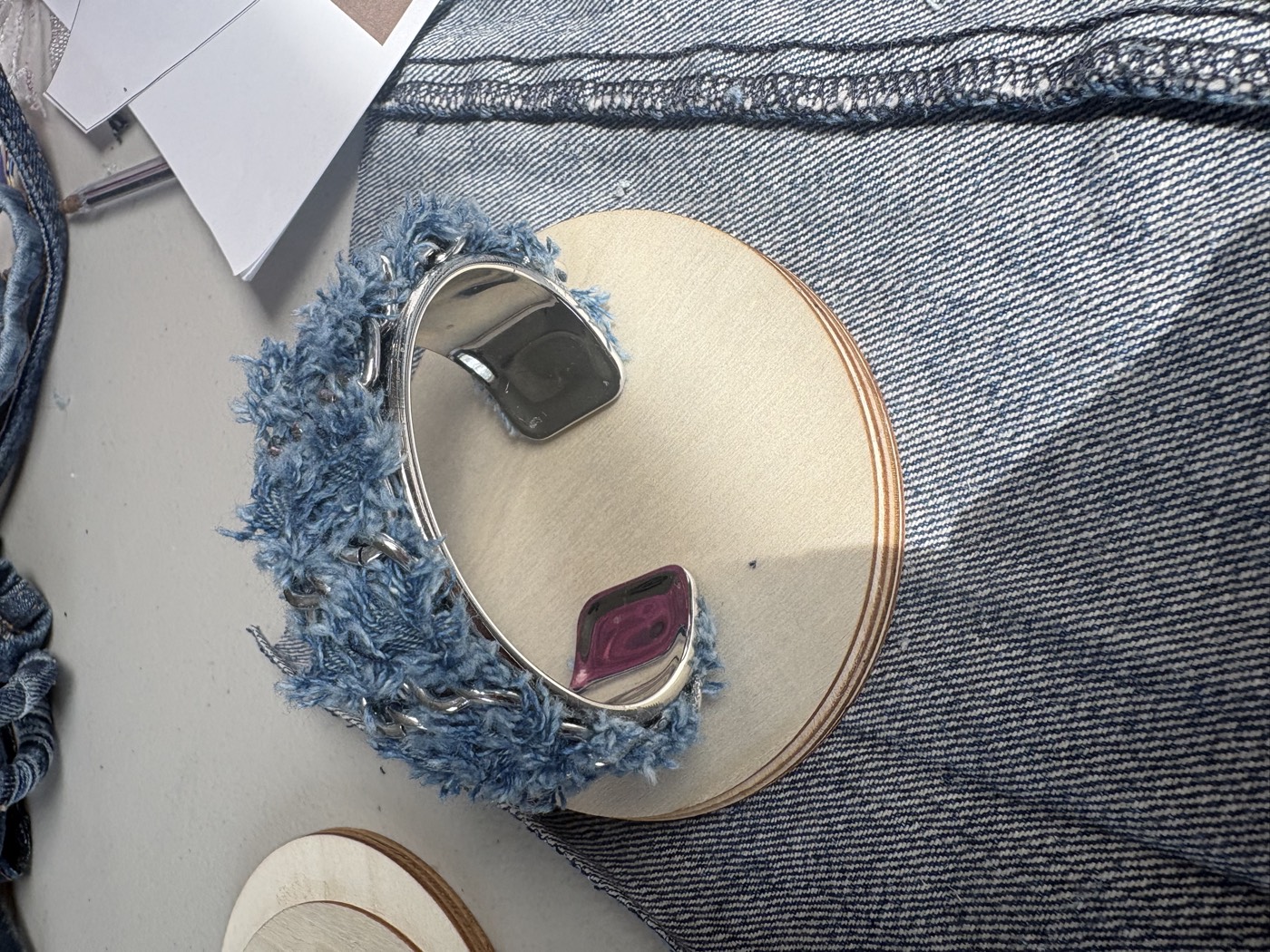 Frayed denim wrapped over a silver cuff bracelet, on a wooden disc on a denim ground.