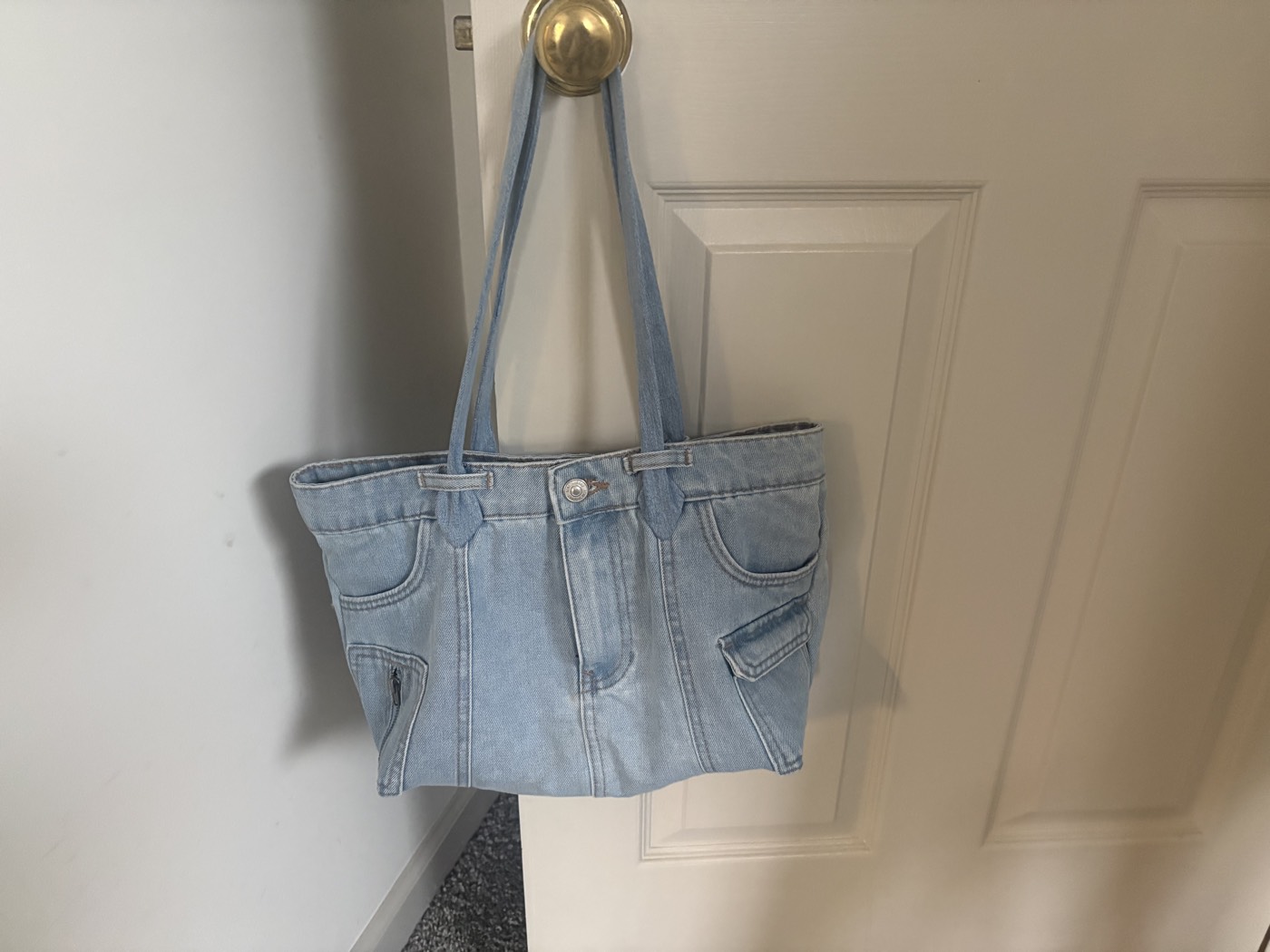 Light-wash denim tote bag with the original waistband, button and pockets, hanging on a door.