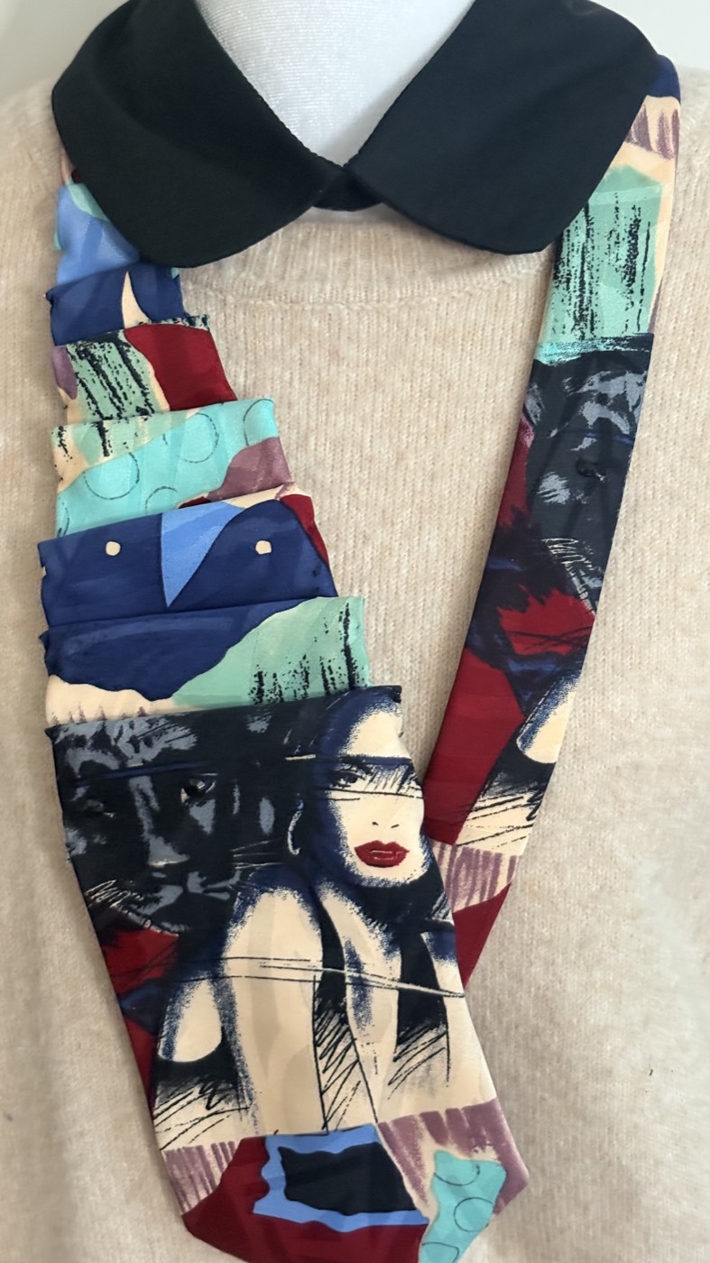 Abstract 1980s print necktie with faces, hands and teal shapes, pleated into a long collar over a dark rounded collar.