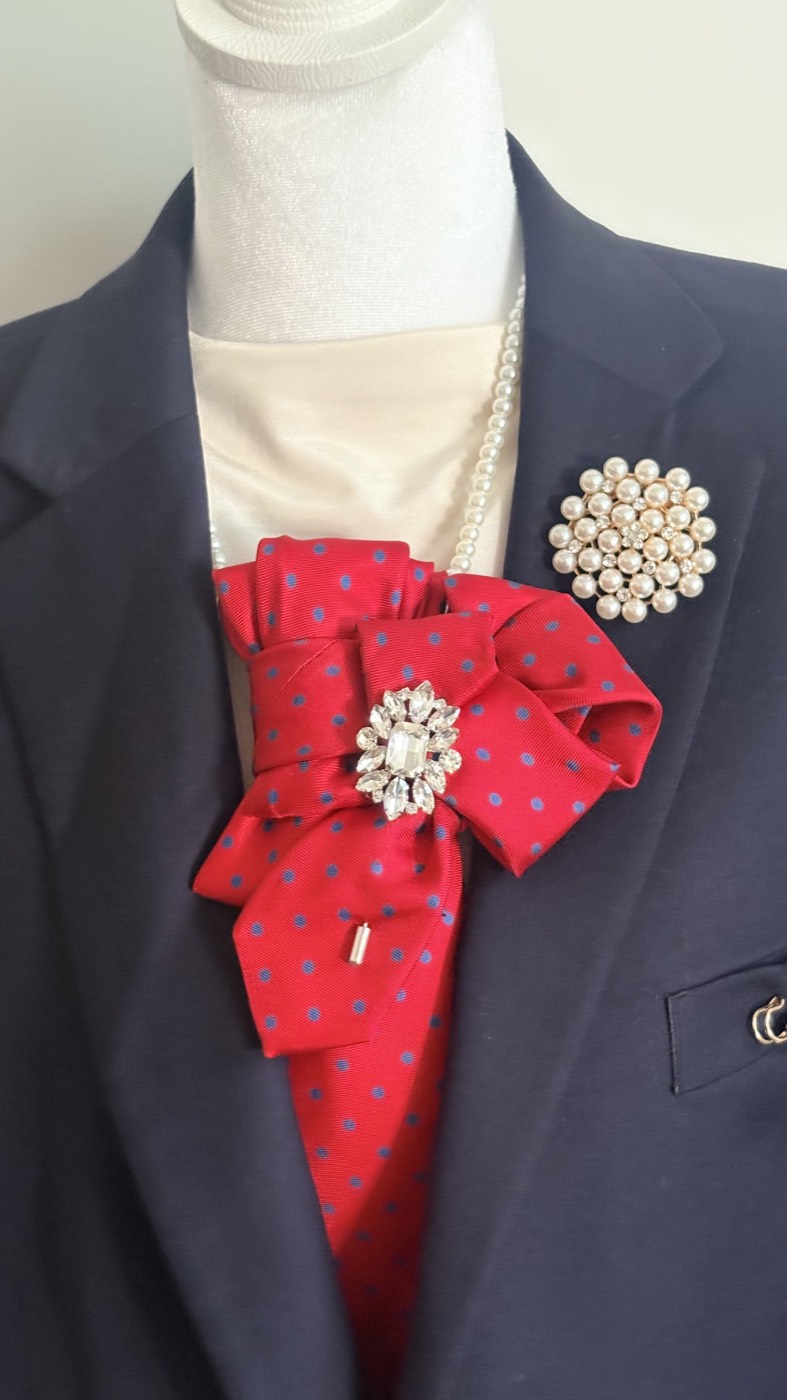 Red necktie with blue polka dots, pleated into a bow with a pearl and crystal brooch, worn over a navy blazer.