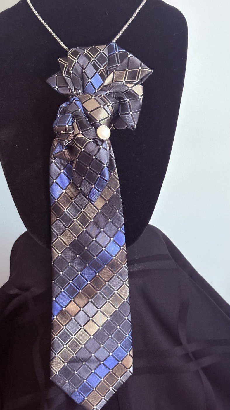 Blue, tan and navy checked necktie knotted into a rosette with a small round brooch, on a silver chain.