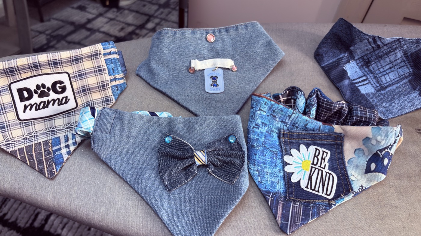 Five patchwork denim dog bandanas laid out on a couch: Dog Mama, Be Kind with a daisy, a bow tie, a dog-face tag and a plaid mix.
