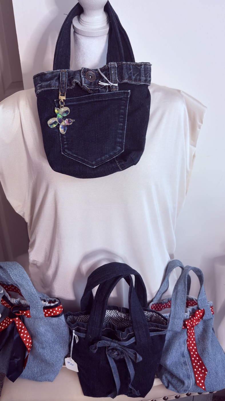 Dark-wash denim tote with a butterfly charm on the zip, on a mannequin, with three mini totes with red polka-dot bows lined up below.