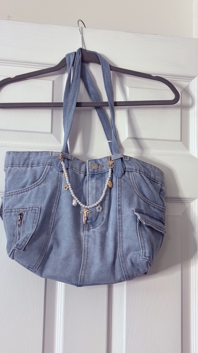 Light-wash denim shoulder bag with two zip pockets and a pearl and charm chain on the strap, hanging on a door.